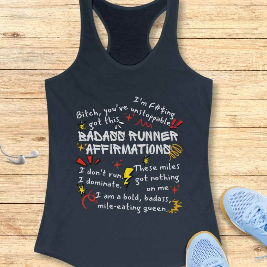 Photograph of a navy blue racerback tank top displaying vibrant 'Badass Runner Affirmations' text in white, red, and yellow, including phrases like 'Bitch, you've got this' and 'I'm f#$ing unstoppable', presented on a light wooden surface with white earbuds and light blue running shoes