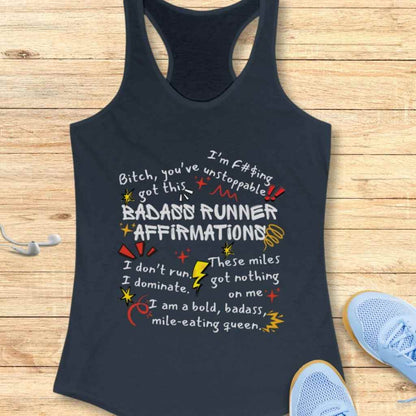 Photograph of a navy blue racerback tank top displaying vibrant 'Badass Runner Affirmations' text in white, red, and yellow, including phrases like 'Bitch, you've got this' and 'I'm f#$ing unstoppable', presented on a light wooden surface with white earbuds and light blue running shoes