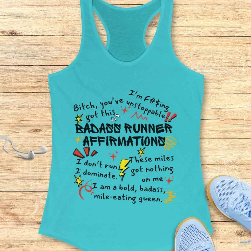 A turquoise racerback tank top featuring colorful bold text of BADASS RUNNER AFFIRMATIONS with various motivational phrases and playful doodle embellishments, arranged on a wooden background next to light blue running shoes and white earbuds