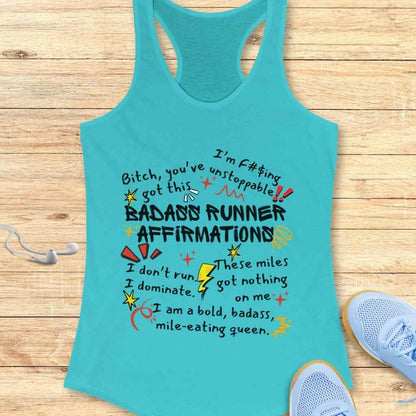 A turquoise racerback tank top featuring colorful bold text of BADASS RUNNER AFFIRMATIONS with various motivational phrases and playful doodle embellishments, arranged on a wooden background next to light blue running shoes and white earbuds