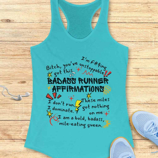 A turquoise racerback tank top featuring colorful bold text of BADASS RUNNER AFFIRMATIONS with various motivational phrases and playful doodle embellishments, arranged on a wooden background next to light blue running shoes and white earbuds