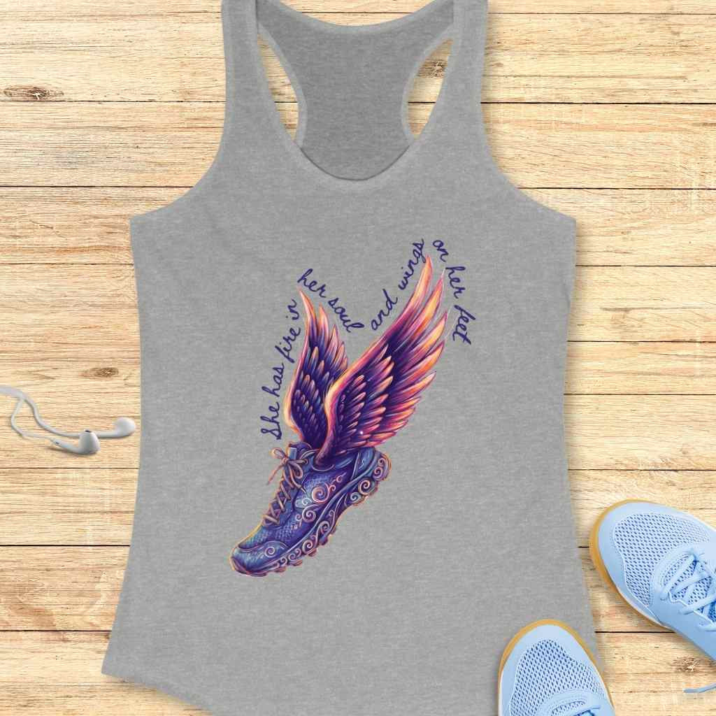 Graphic design on a grey tank top featuring a dark purple running shoe with intricate scrollwork and vibrant purple, orange, and pink gradient wings, accompanied by the inspirational text "She has fire in her soul and wings on her feet" set against a light wooden background with white earbuds and light blue running shoes