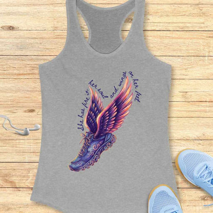 Graphic design on a grey tank top featuring a dark purple running shoe with intricate scrollwork and vibrant purple, orange, and pink gradient wings, accompanied by the inspirational text "She has fire in her soul and wings on her feet" set against a light wooden background with white earbuds and light blue running shoes