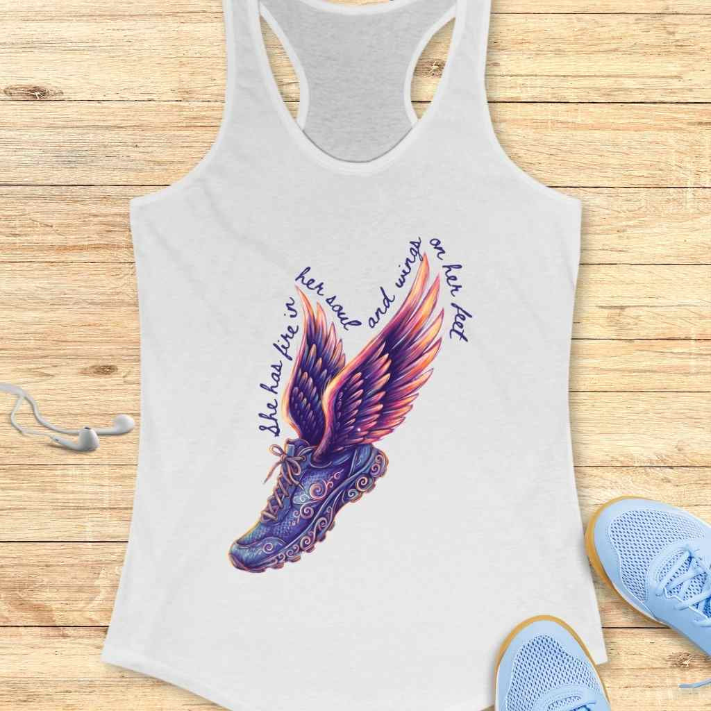 White racerback tank top displaying a vibrant graphic of a purple and orange winged running shoe accompanied by the text 'She has fire in her soul and wings on her feet' placed on a light wood surface alongside blue sneakers and white earbuds