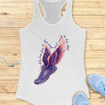 White racerback tank top displaying a vibrant graphic of a purple and orange winged running shoe accompanied by the text 'She has fire in her soul and wings on her feet' placed on a light wood surface alongside blue sneakers and white earbuds