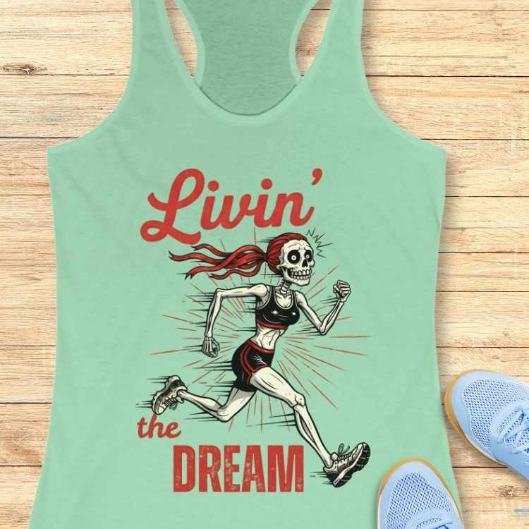 Graphic design on a mint green racerback tank top featuring a cartoonish female skeleton runner with a vibrant red ponytail, wearing a black sports bra and shorts, a red sunburst effect, and text Livin' the DREAM, all placed near light blue running shoes on a wooden surface
