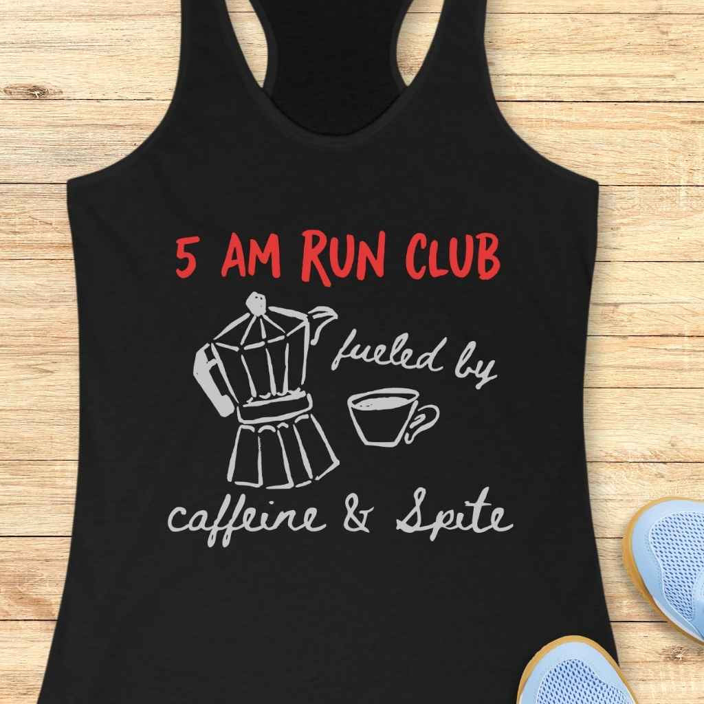 Photo of a black tank top displaying the red text 5 AM RUN CLUB and white text fueled by caffeine & Spite with a white line drawing of a moka pot and coffee cup, laid flat on a light wood background with light blue athletic shoes partially visible