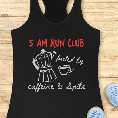 Photo of a black tank top displaying the red text 5 AM RUN CLUB and white text fueled by caffeine & Spite with a white line drawing of a moka pot and coffee cup, laid flat on a light wood background with light blue athletic shoes partially visible