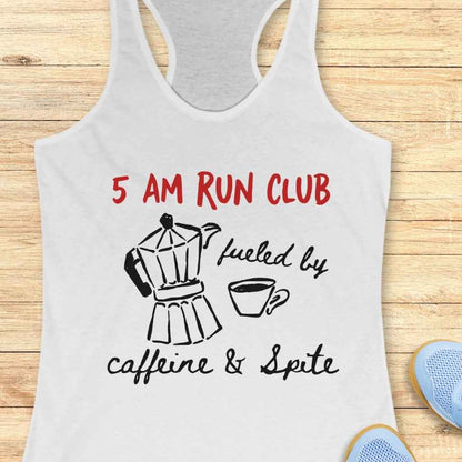 A white graphic tank top featuring the red text "5 AM RUN CLUB" and black script "fueled by caffeine & Spite", accompanied by a black line art Moka pot and coffee cup illustration, displayed on a wooden background next to blue running shoes