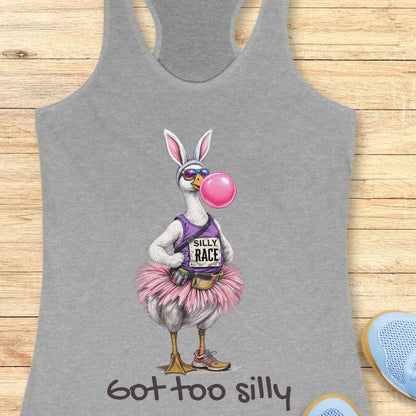 Digital illustration printed on a grey tank top showcasing a quirky goose character wearing white bunny ears, purple sunglasses, and blowing a vibrant pink bubblegum bubble, dressed in a purple 'Silly Race' tank top, a fluffy pink tutu, and tiny running shoes, with the phrase 'got too silly' printed below, displayed against a light wooden background with blue sneakers in the bottom right corner