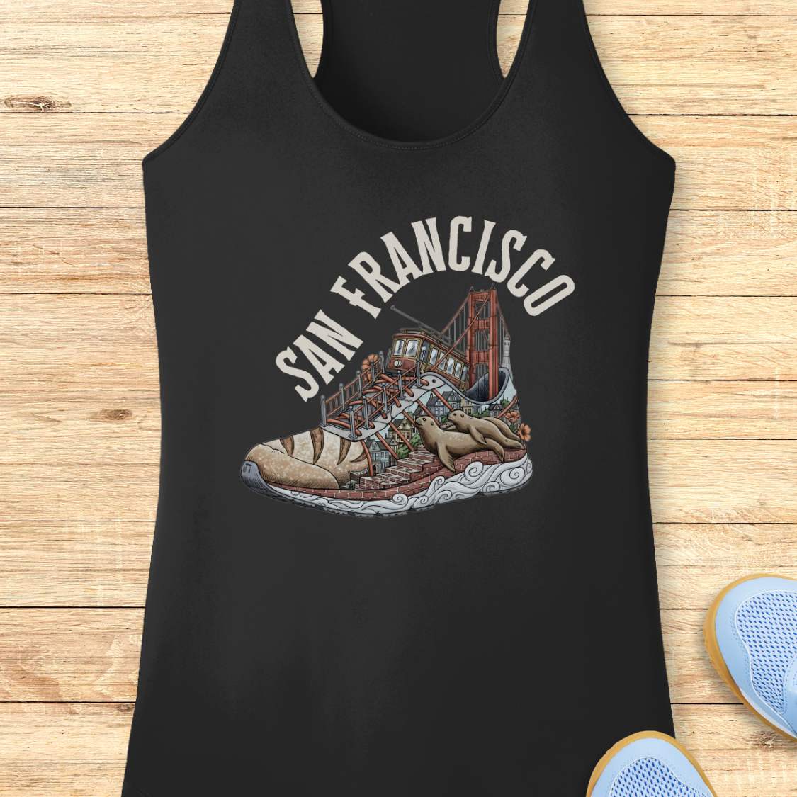 A black racerback tank top laid flat on a light wood background, featuring a distressed white 'SAN FRANCISCO' graphic above an illustrated sneaker with a bridge motif. Part of a blue shoe is visible in the bottom right corner.