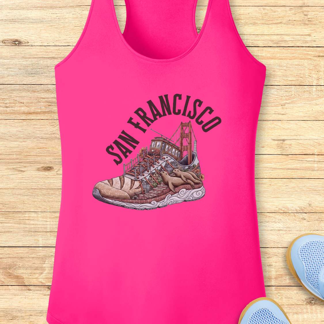 A bright pink racerback tank top laid flat on a light wood plank background, featuring a graphic of a sneaker with the Golden Gate Bridge and the words 'SAN FRANCISCO' in arched black text.