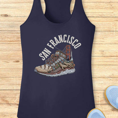A navy blue racerback tank top laid flat on a light wood plank background, featuring a graphic print of a sneaker with the Golden Gate Bridge and the words 'SAN FRANCISCO' in white arched text above it. Part of a light blue shoe is visible in the bottom right corner as a styling prop.