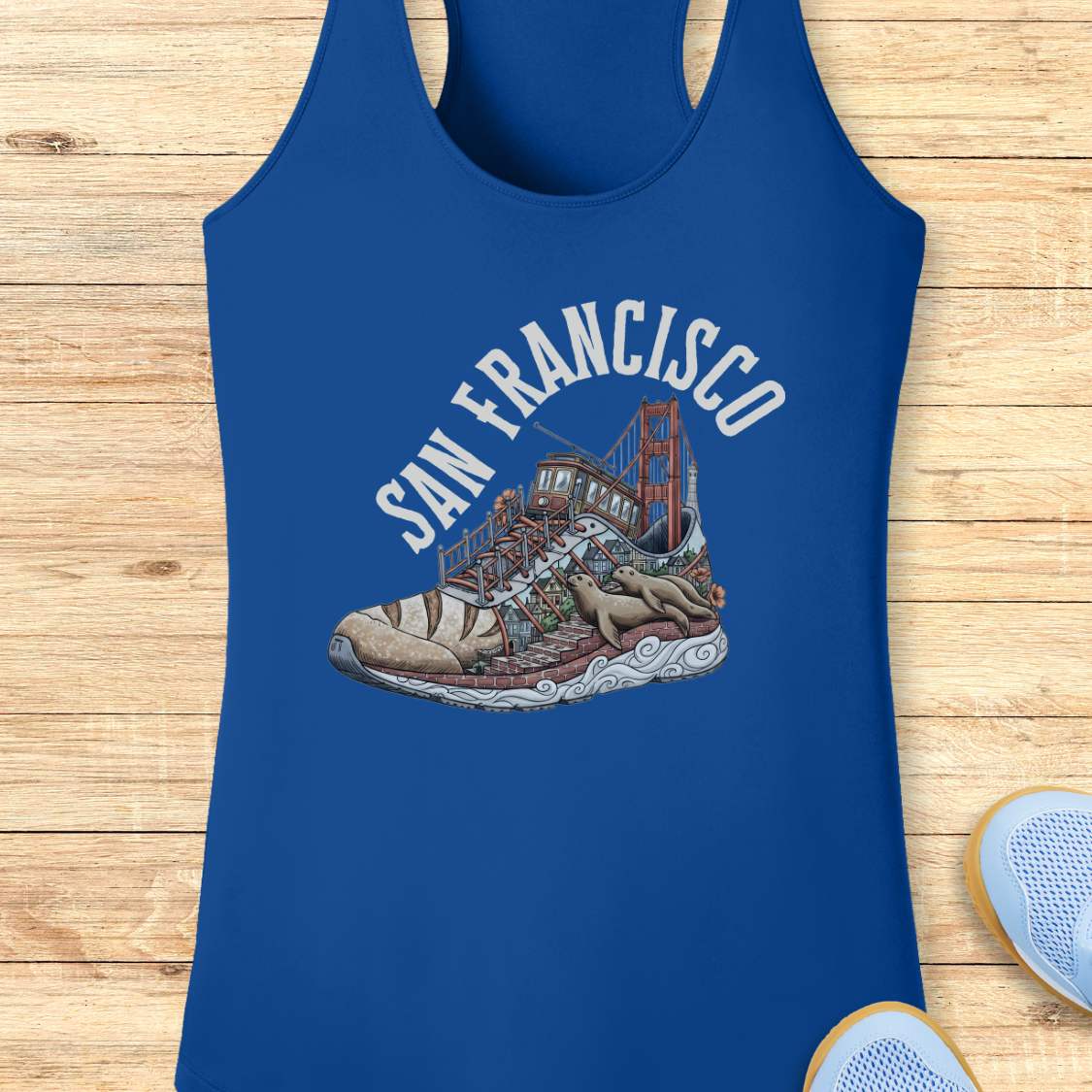 A royal blue racerback tank top laid flat on a light wood background, featuring a graphic print of a sneaker with the Golden Gate Bridge and the words 'SAN FRANCISCO' in white arched text.