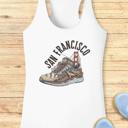 A white racerback tank top laid flat on a light wood background, featuring a graphic print of a sneaker with the Golden Gate Bridge and the words 'SAN FRANCISCO' in arched black text.