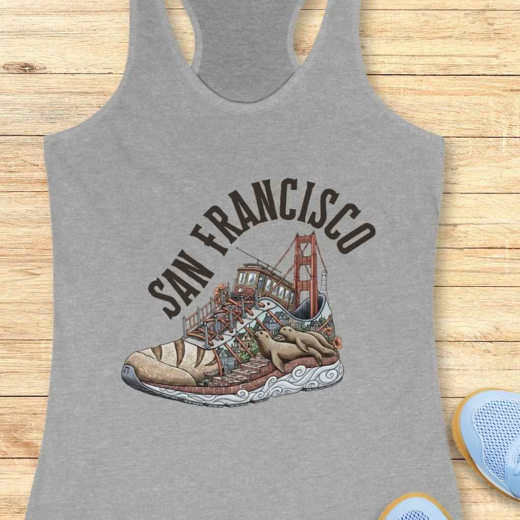 Screen-printed graphic on a grey tank top showcasing a stylized running shoe filled with a vibrant San Francisco cityscape featuring the Golden Gate Bridge, a classic cable car, brick streets, and two lounging sea lions, all beneath the arched text SAN FRANCISCO