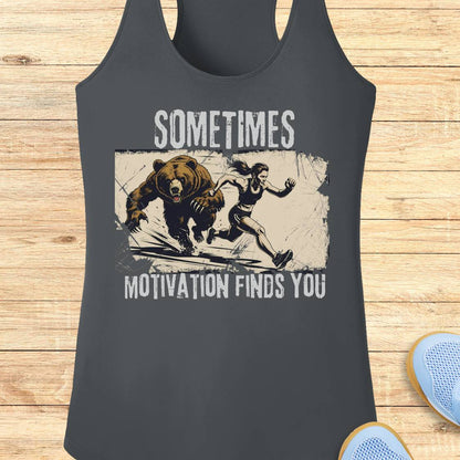 A dark gray racerback tank top with a graphic print of a woman running from a bear, with the text 'SOMETIMES MOTIVATION FINDS YOU' in distressed white lettering. The tank top is displayed flat on a light wood background with blue sneakers partially visible in the corner.