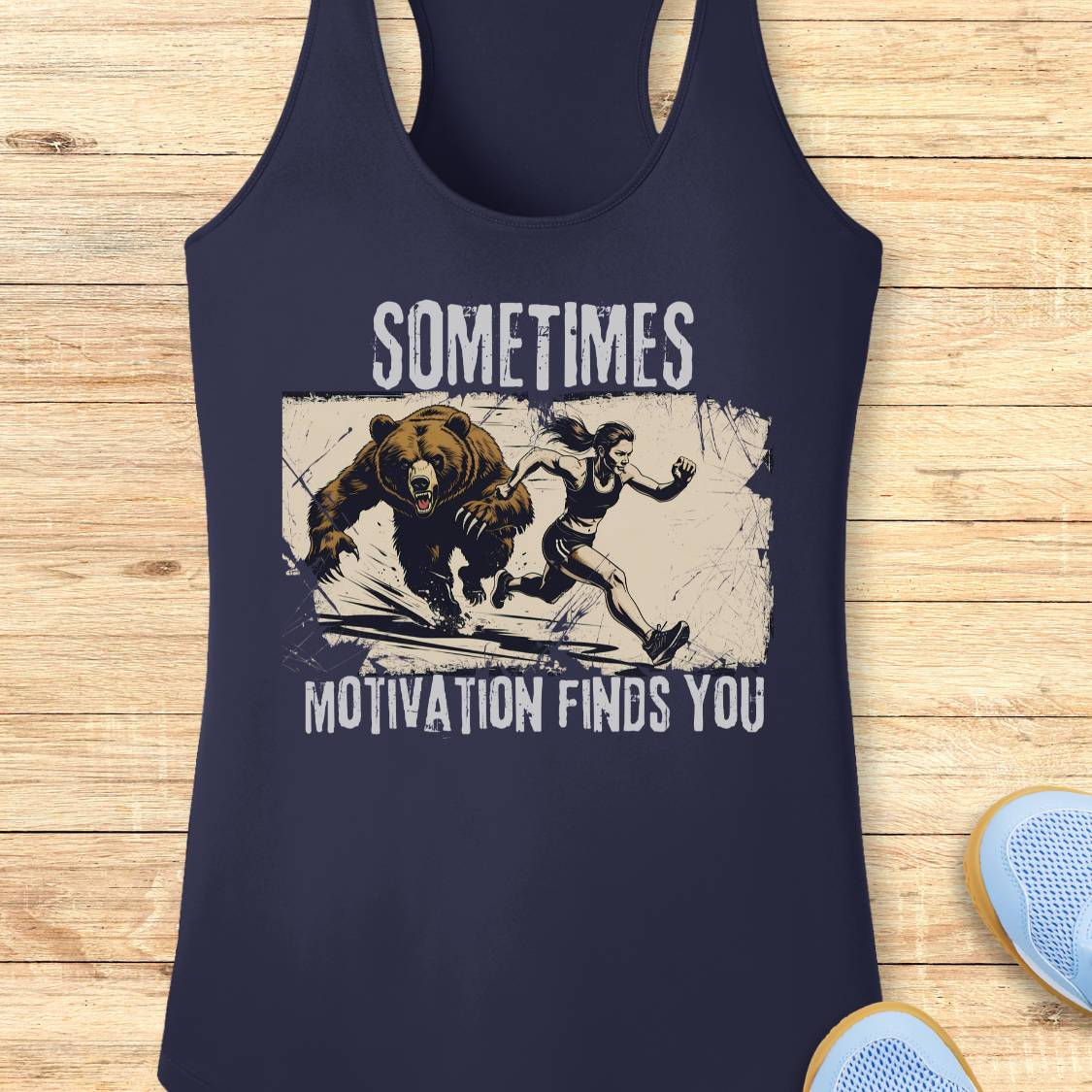 A navy blue racerback tank top with a distressed graphic of a bear chasing a runner, with the text 'SOMETIMES MOTIVATION FINDS YOU' printed in white. The tank top is displayed flat on a light wood background with a pair of blue sneakers partially visible in the corner.