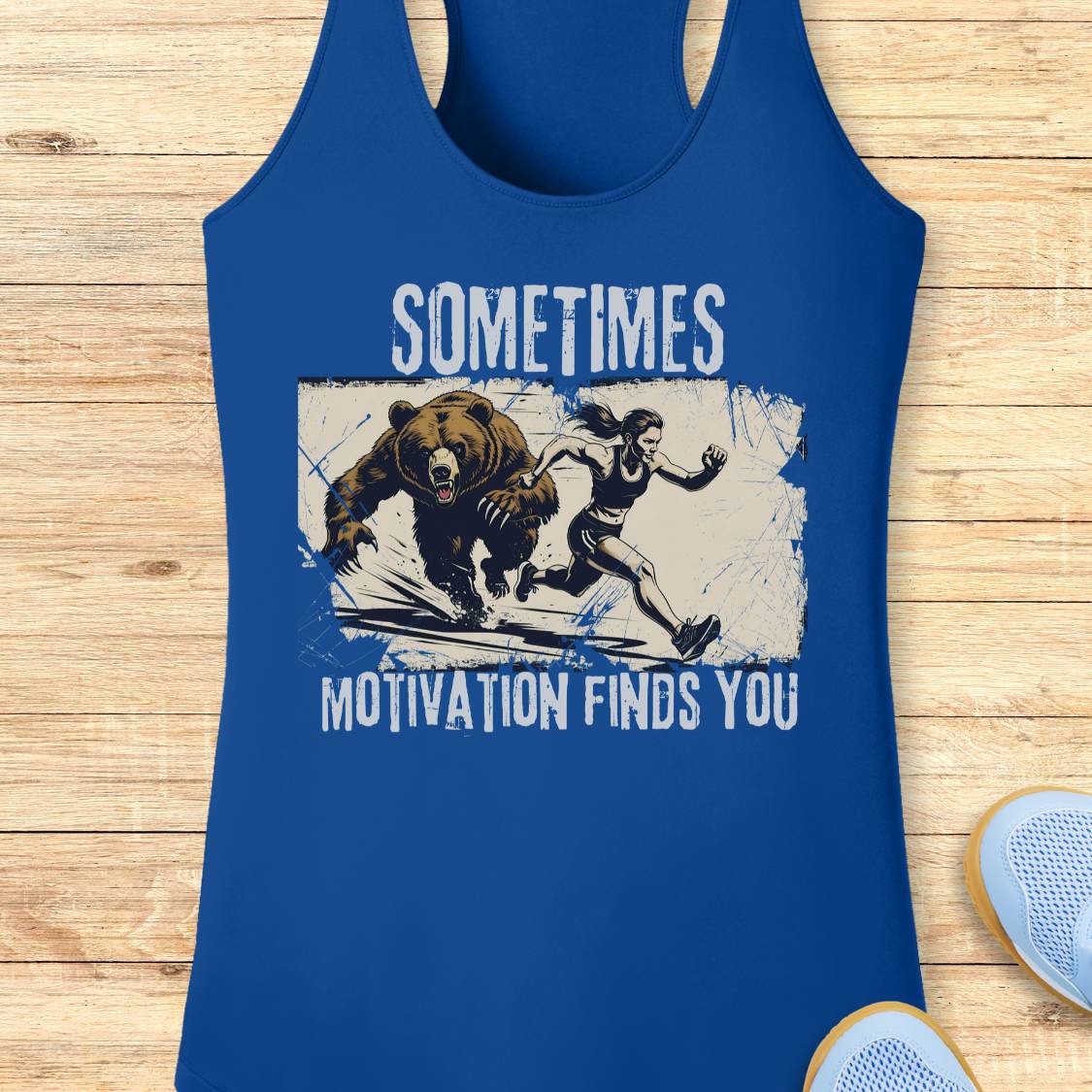 A royal blue racerback tank top with a graphic print of a woman running from a bear, featuring the text 'SOMETIMES MOTIVATION FINDS YOU' in white. The tank top is displayed flat on a light wood background with a sneaker partially visible in the corner.
