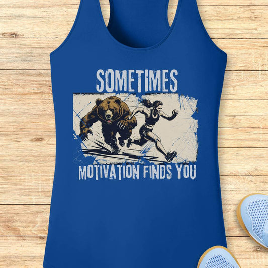 A royal blue racerback tank top with a graphic print of a woman running from a bear, featuring the text 'SOMETIMES MOTIVATION FINDS YOU' in white. The tank top is displayed flat on a light wood background with a sneaker partially visible in the corner.