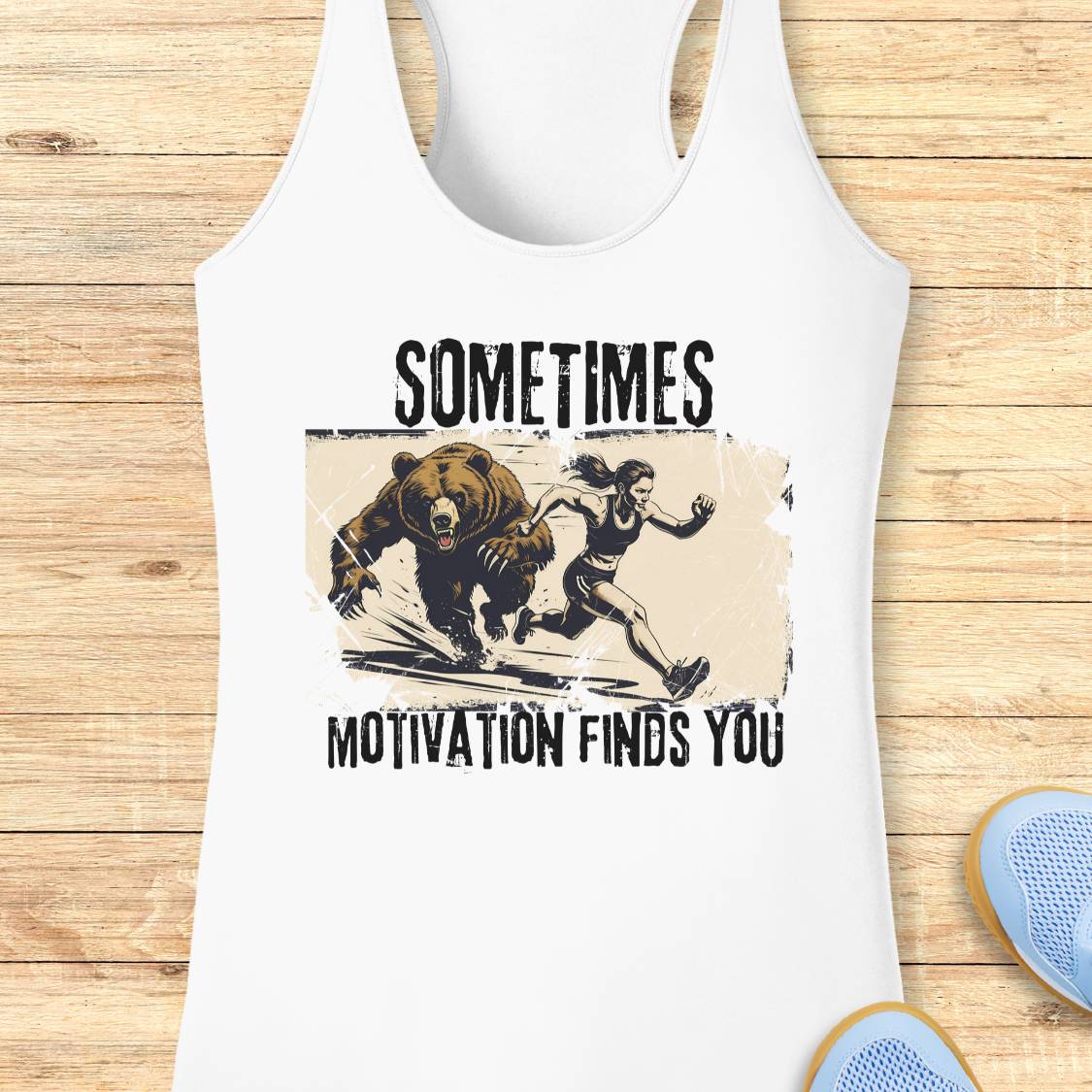 White racerback tank top laid flat on a light wood background, featuring a distressed graphic of a woman running from a bear with the text 'SOMETIMES MOTIVATION FINDS YOU'.