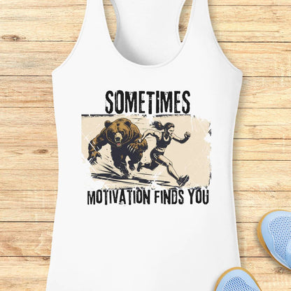 White racerback tank top laid flat on a light wood background, featuring a distressed graphic of a woman running from a bear with the text 'SOMETIMES MOTIVATION FINDS YOU'.