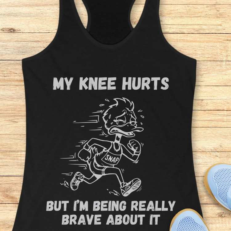 Graphic design on a black tank top featuring a distressed cartoon duck runner wearing a "SNAP" bib with white text "MY KNEE HURTS BUT I'M BEING REALLY BRAVE ABOUT IT" displayed on a wooden surface beside light blue sneakers