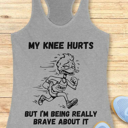 Graphic design on a grey tank top featuring a cartoon duck crying and sweating in a 'SNAP' bib while running, with the text MY KNEE HURTS BUT I'M BEING REALLY BRAVE ABOUT IT