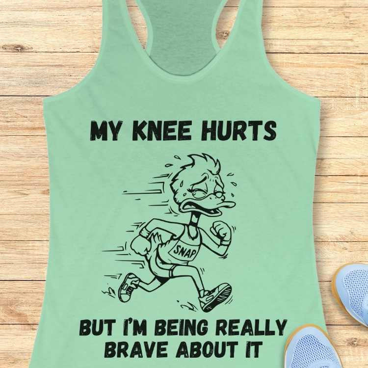 Graphic print on a light green tank top featuring a pained cartoon duck running wearing a "SNAP" shirt and the text "MY KNEE HURTS BUT I'M BEING REALLY BRAVE ABOUT IT" displayed on a wooden surface next to light blue running shoes