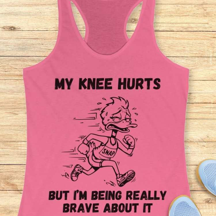 Printed graphic on a pink racerback tank top featuring a sweating, grimacing cartoon duck runner wearing a "SNAP" bib and the text "MY KNEE HURTS BUT I'M BEING REALLY BRAVE ABOUT IT" displayed on a wooden plank background next to light blue running shoes