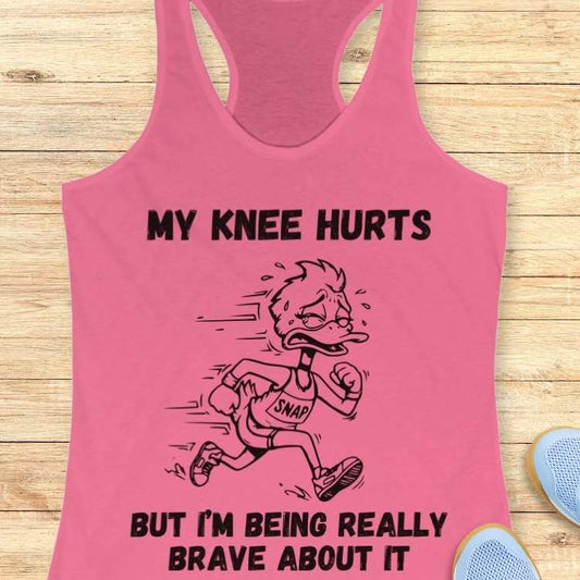 Printed graphic on a pink racerback tank top featuring a sweating, grimacing cartoon duck runner wearing a "SNAP" bib and the text "MY KNEE HURTS BUT I'M BEING REALLY BRAVE ABOUT IT" displayed on a wooden plank background next to light blue running shoes