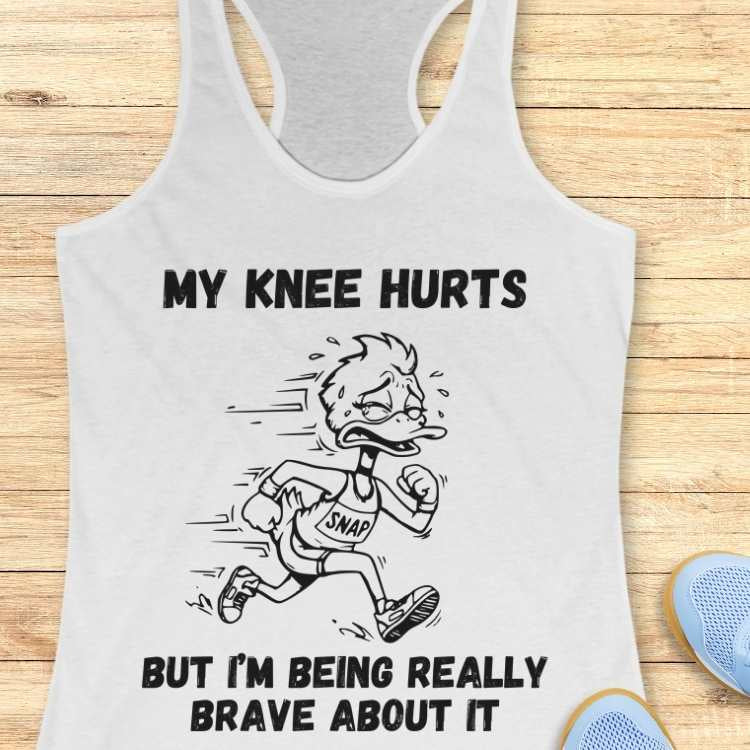 Graphic design on a white tank top featuring a black and white cartoon duck running with a pained grimace and sweat droplets, wearing a "SNAP" race bib, with the bold text "MY KNEE HURTS BUT I'M BEING REALLY BRAVE ABOUT IT" and light blue running shoes partially visible on a wooden background