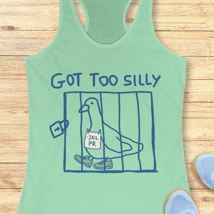 Graphic design on a mint green tank top showing a blue outline drawing of a pigeon in a cage wearing sneakers with a -JAIL PR sign around its neck and the text GOT TOO SILLY above, displayed against a wooden background with light blue sneakers visible