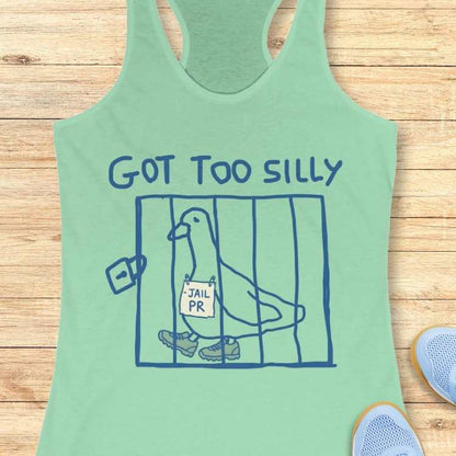 Graphic design on a mint green tank top showing a blue outline drawing of a pigeon in a cage wearing sneakers with a -JAIL PR sign around its neck and the text GOT TOO SILLY above, displayed against a wooden background with light blue sneakers visible