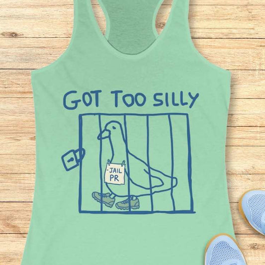 Graphic design on a mint green tank top showing a blue outline drawing of a pigeon in a cage wearing sneakers with a -JAIL PR sign around its neck and the text GOT TOO SILLY above, displayed against a wooden background with light blue sneakers visible