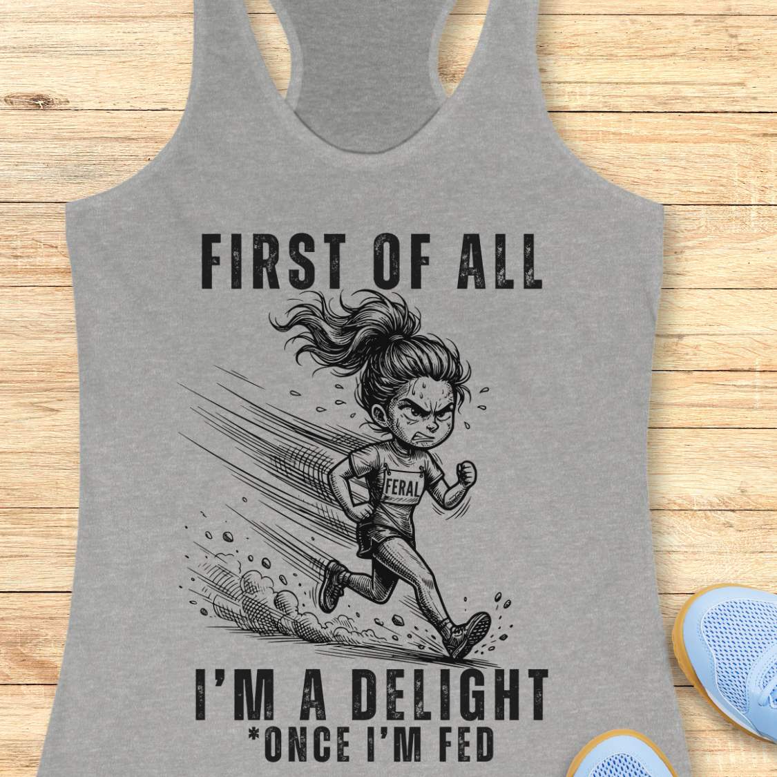 Gray tank top with graphic of a running girl and text first  of all I'm a delight , once I'm fed, on a wooden surface