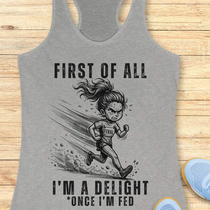 Gray tank top with graphic of a running girl and text first  of all I'm a delight , once I'm fed, on a wooden surface