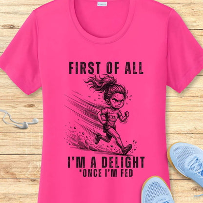 pink performance tee with a graphic of a running girl and text first  of all I'm a delight , once I'm fed, on a wooden surface