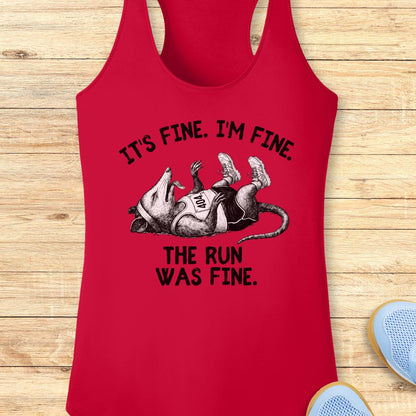 A red racerback performance tank top laid flat on a light wood background, featuring a black graphic of a running mouse with the text 'IT'S FINE. I'M FINE.
