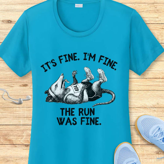 blue performance t-shirt with possum image and text 'It's fine. I'm fine. The run was fine.'  on a wooden surface with sneakers.