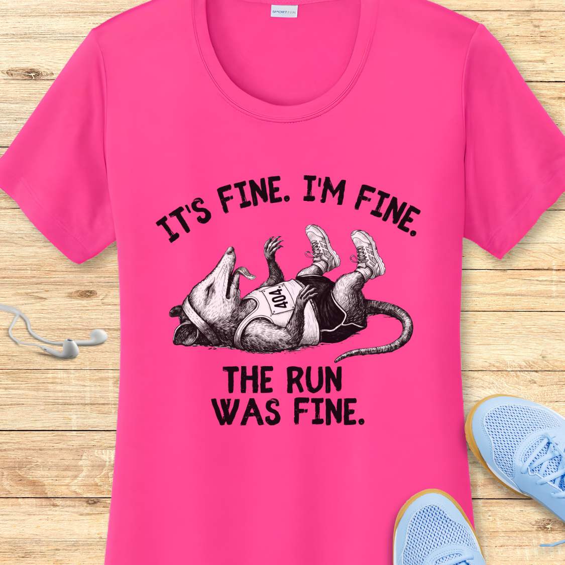 pink performance t-shirt with possum image and text 'It's fine. I'm fine. The run was fine.'  on a wooden surface with sneakers.