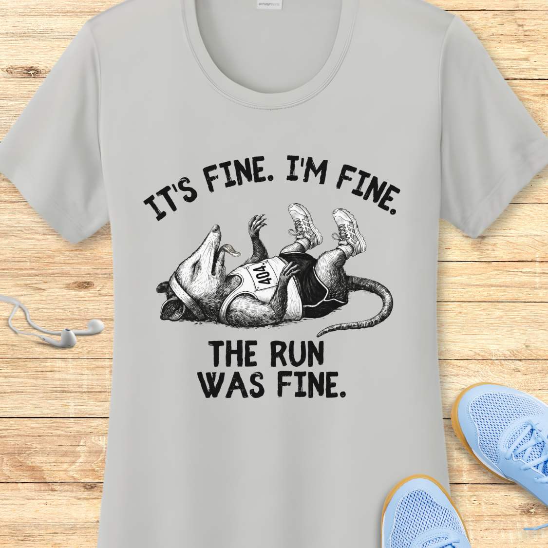 silver performance t-shirt with possum image and text 'It's fine. I'm fine. The run was fine.'  on a wooden surface with sneakers.