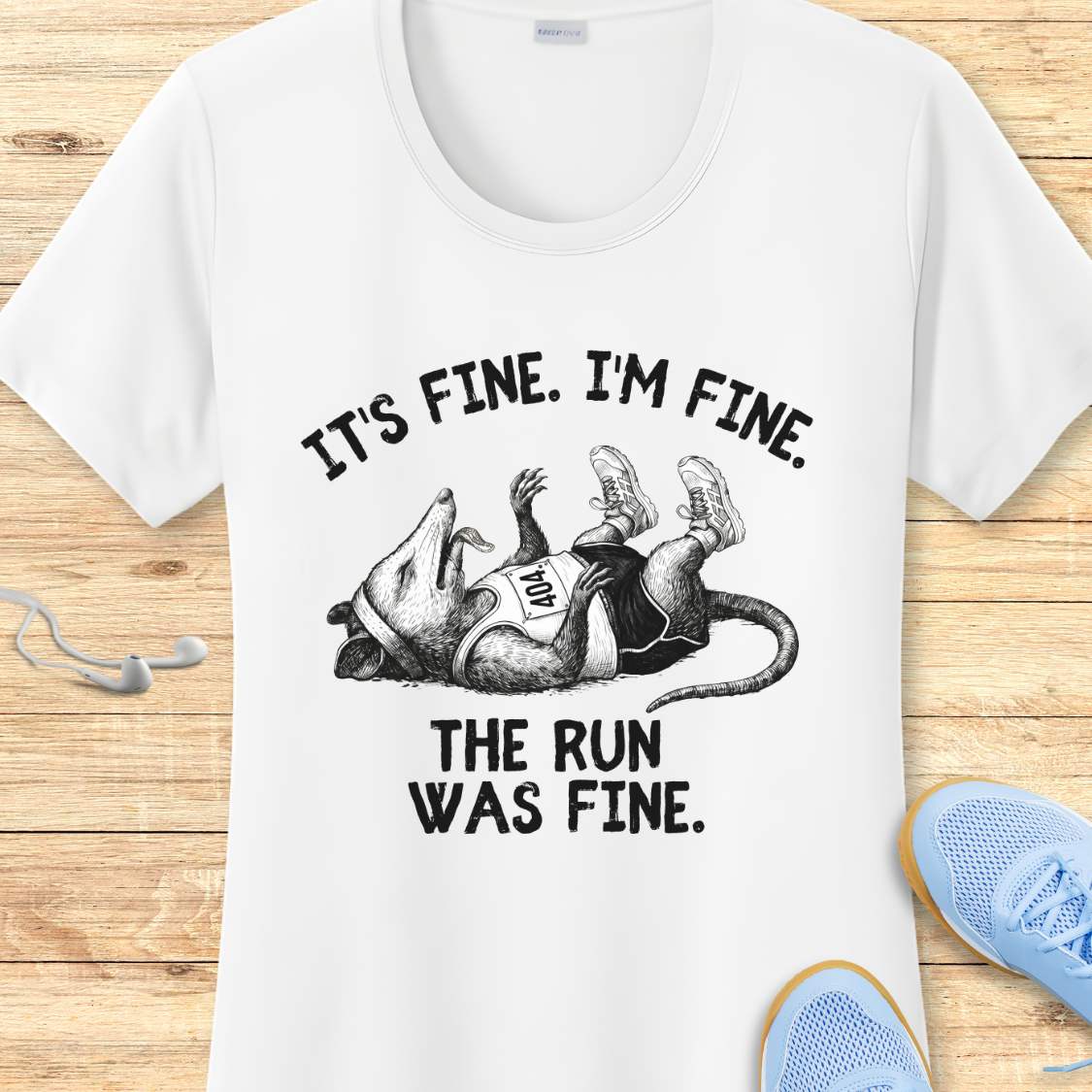 white performance t-shirt with possum image and text 'It's fine. I'm fine. The run was fine.'  on a wooden surface with sneakers.