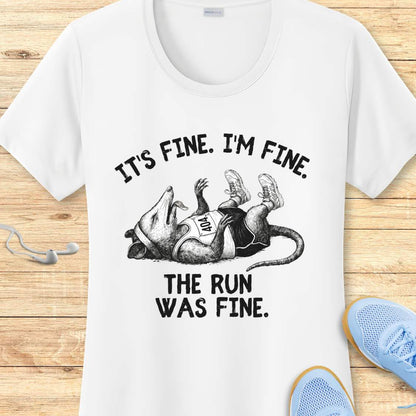 white performance t-shirt with possum image and text 'It's fine. I'm fine. The run was fine.'  on a wooden surface with sneakers.