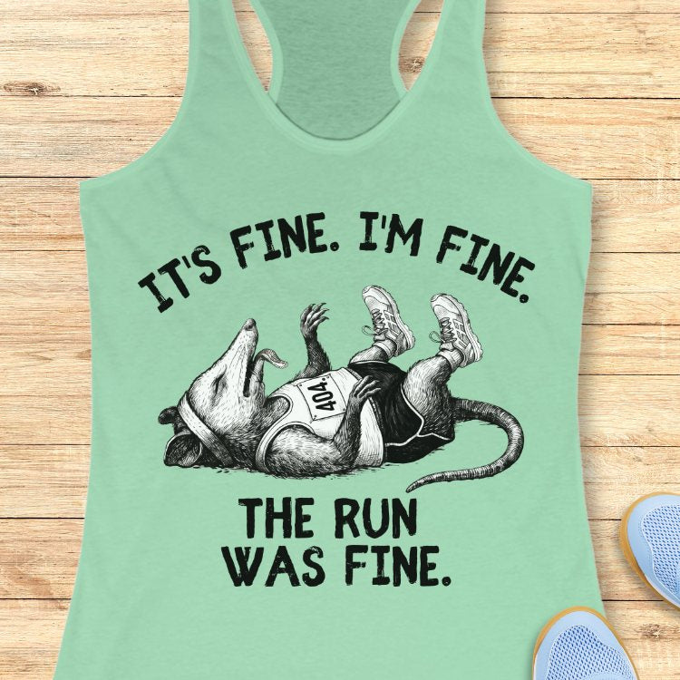 Mint green tank top with graphic of a possum and text 'It's fine. I'm fine. The run was fine.' on a wooden surface.