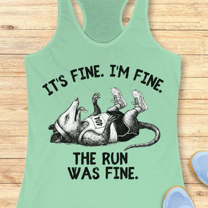 Mint green tank top with graphic of a possum and text 'It's fine. I'm fine. The run was fine.' on a wooden surface.