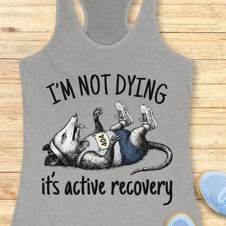 Gray tank top with a graphic of a rat and text 'I'm not dying, it's active recovery' on a wooden surface.
