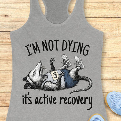 Gray tank top with a graphic of a rat and text 'I'm not dying, it's active recovery' on a wooden surface.