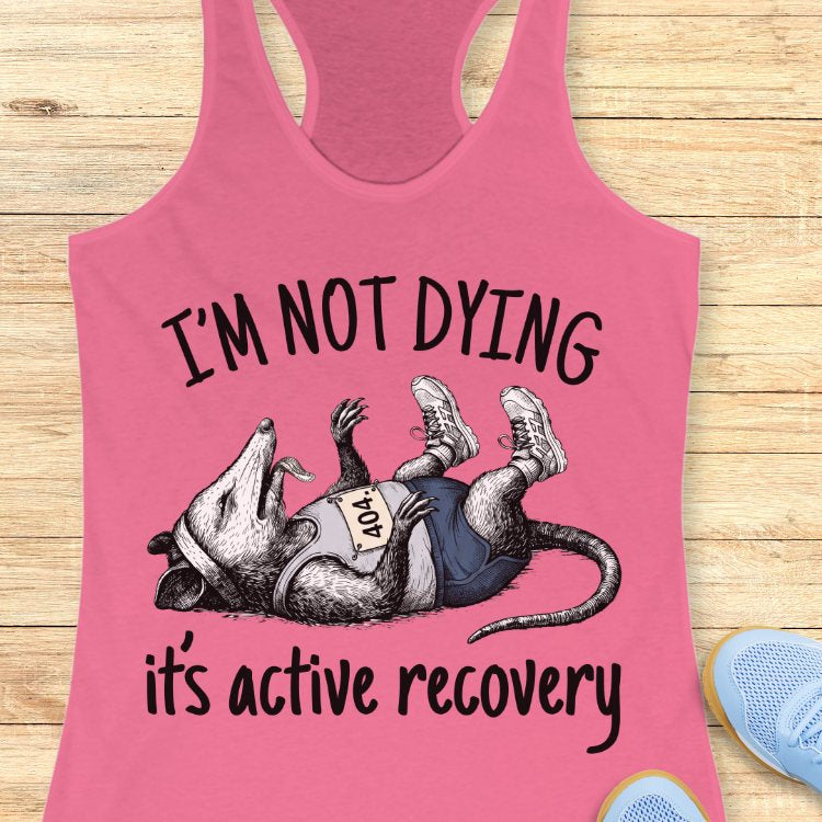 Pink tank top with possum graphic and text 'I'm not dying, it's active recovery' on a wooden surface.