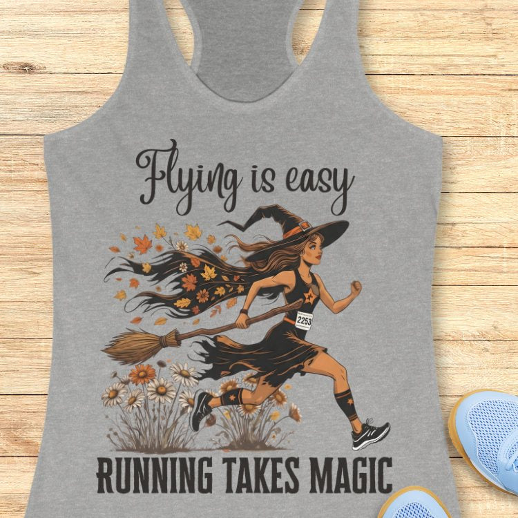 Gray tank top with a witch running on a broom design and text 'Flying is easy, running takes magic' on a wooden surface.